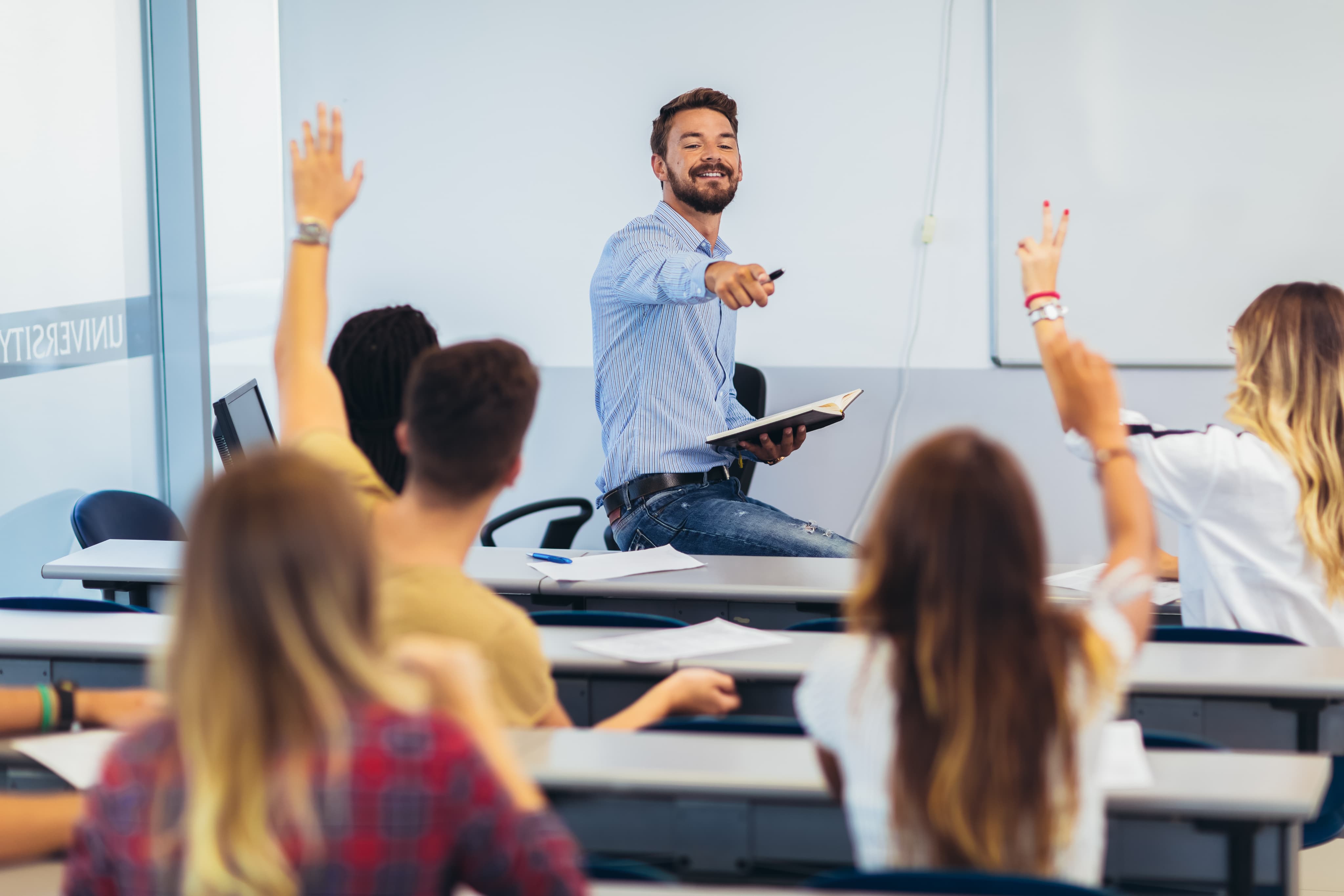 A teacher sitting on their desk pointing to their students.