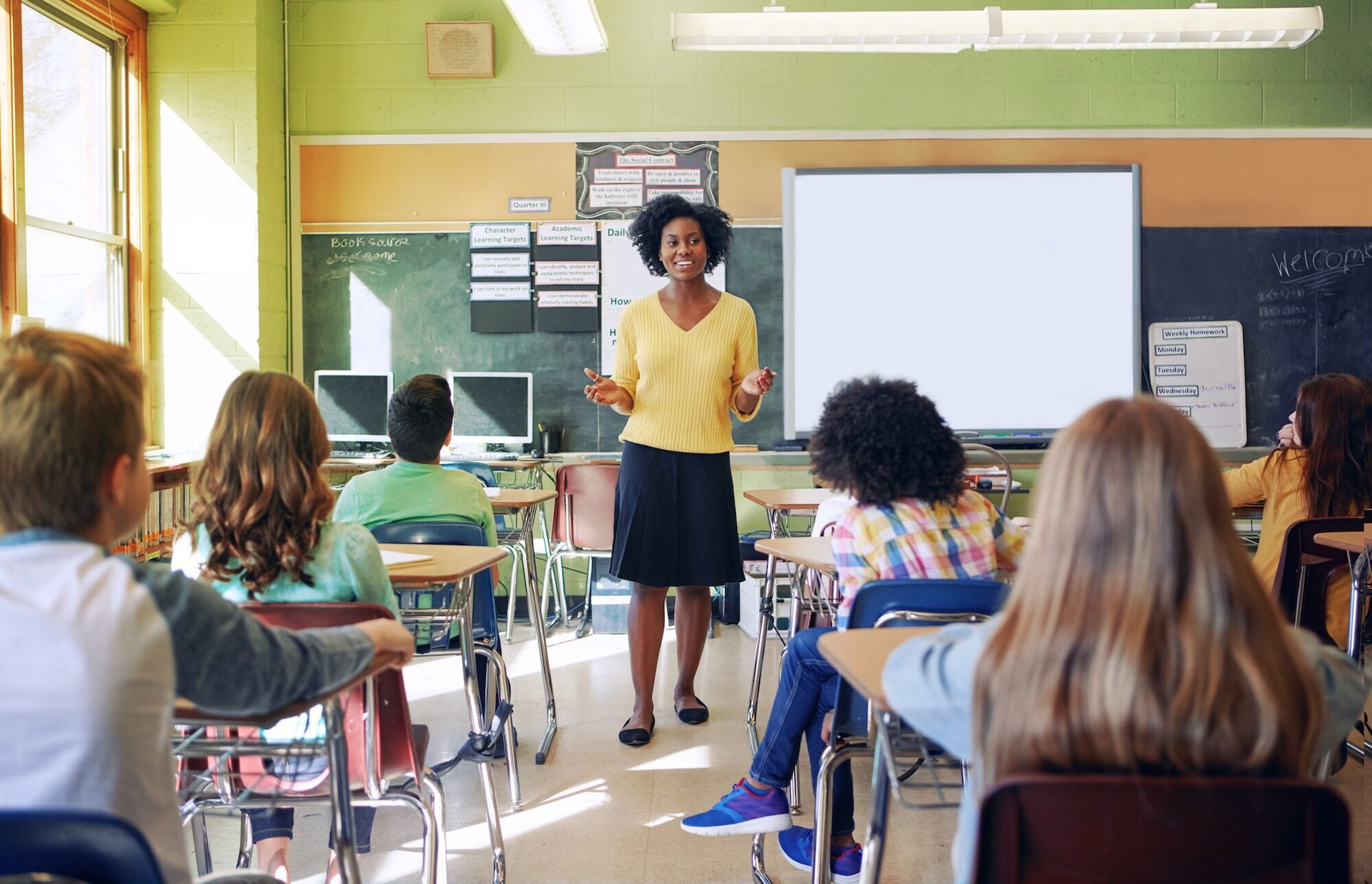 A teacher standing in front of a class of children sitting in desks.
